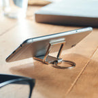 A silver metal phone stand keychain holding a smartphone on a wooden desk, with a blurred laptop and glasses in the background.