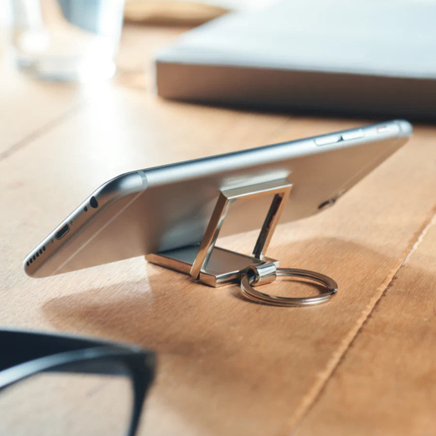 A silver metal phone stand keychain holding a smartphone on a wooden desk, with a blurred laptop and glasses in the background.