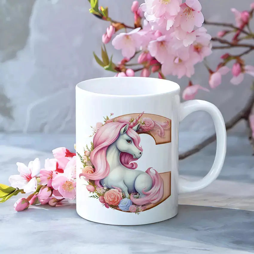 A white ceramic mug featuring a printed illustration of a white unicorn with pink hair and a gold letter 'C' background, styled on a gray marble surface with pink cherry blossoms.