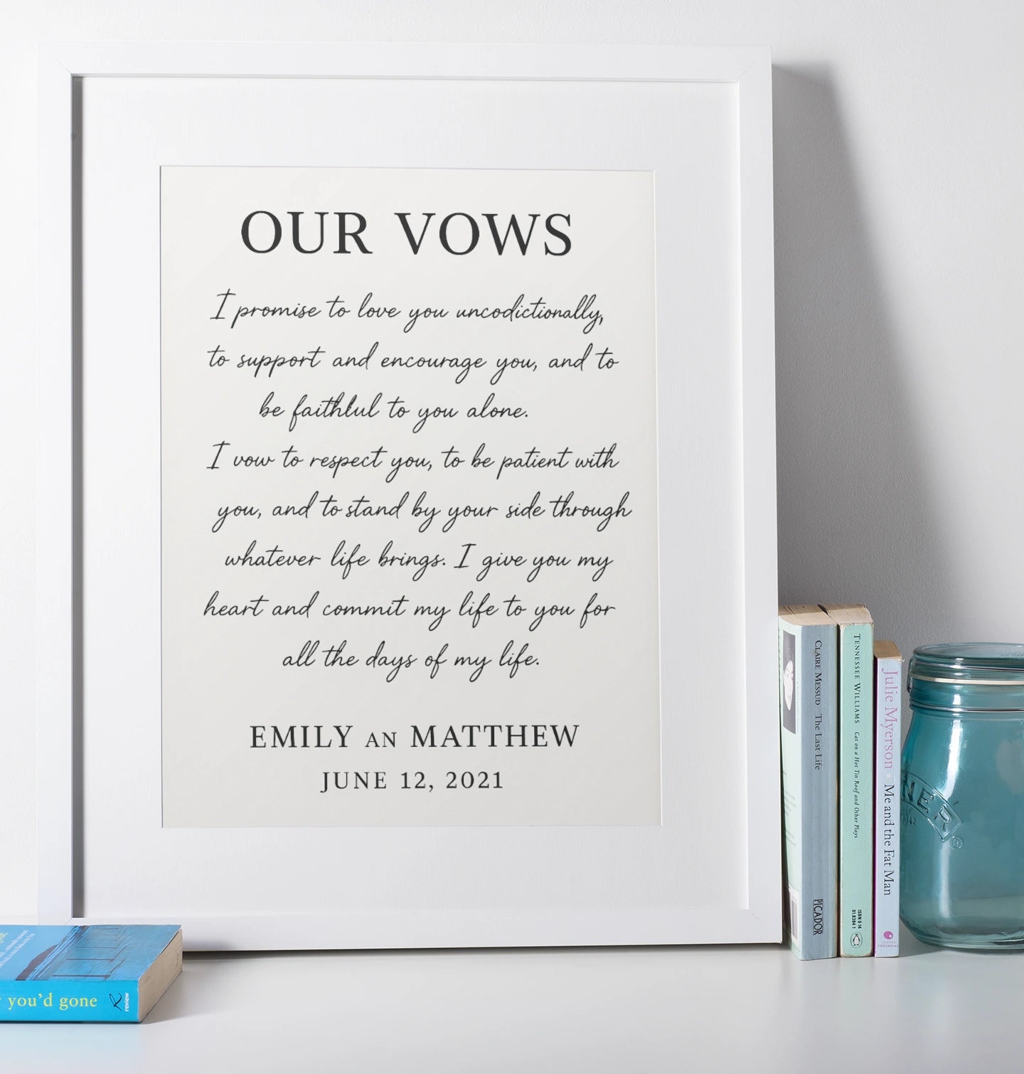 Framed print of wedding vows on a white surface with books and a jar.