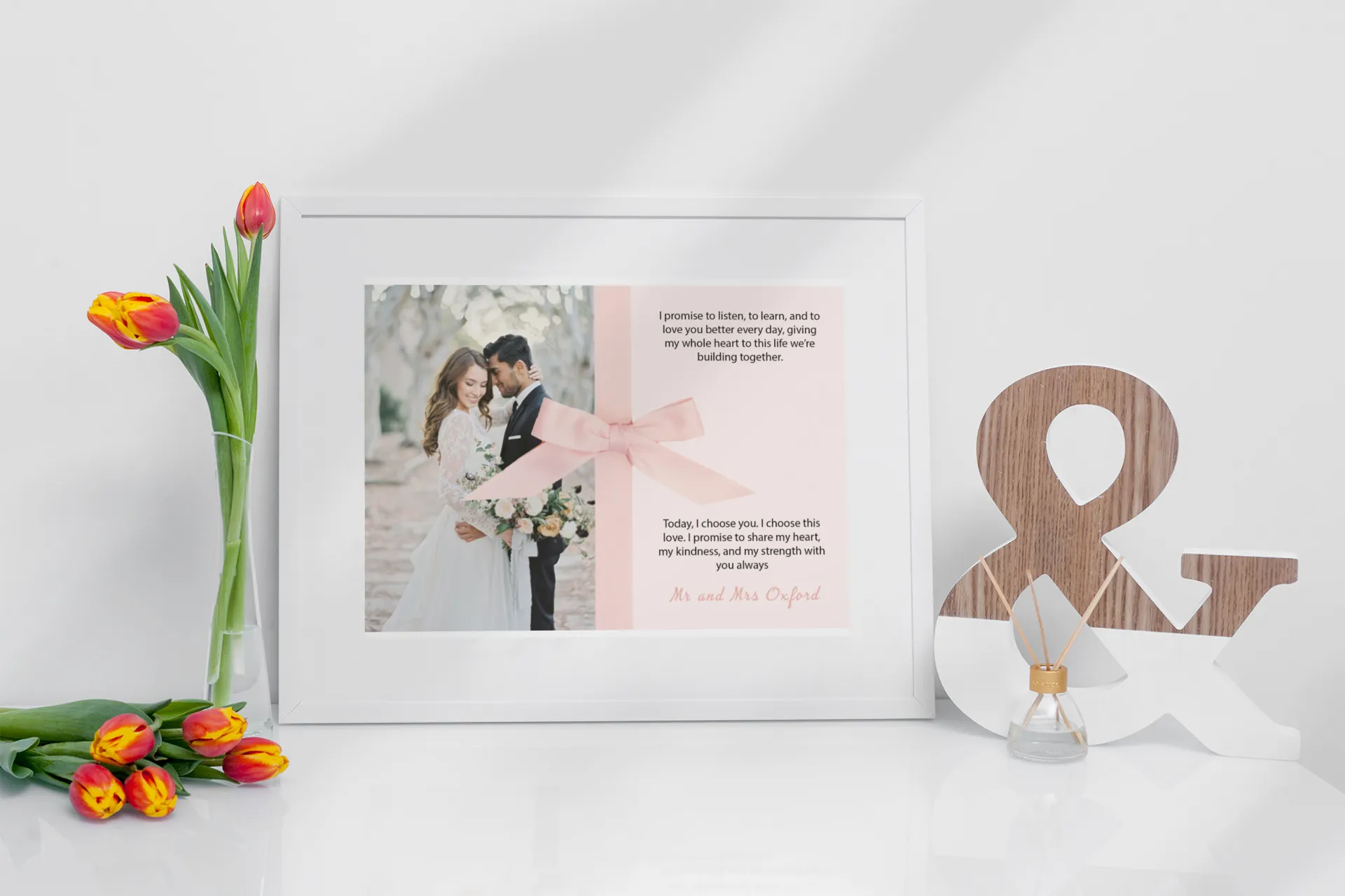 “Personalised wedding vow print with couple’s photo in a white frame, featuring a pink ribbon design, displayed on a white tabletop with tulips and décor.”